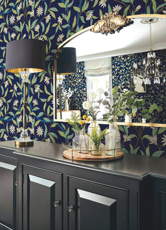 A room adorned with Jasmine Wallpaper by York Wallcoverings features a gold-framed mirror, black lamp, chandelier, dark cabinet, and a tray displaying glass vases filled with flowers and greenery.