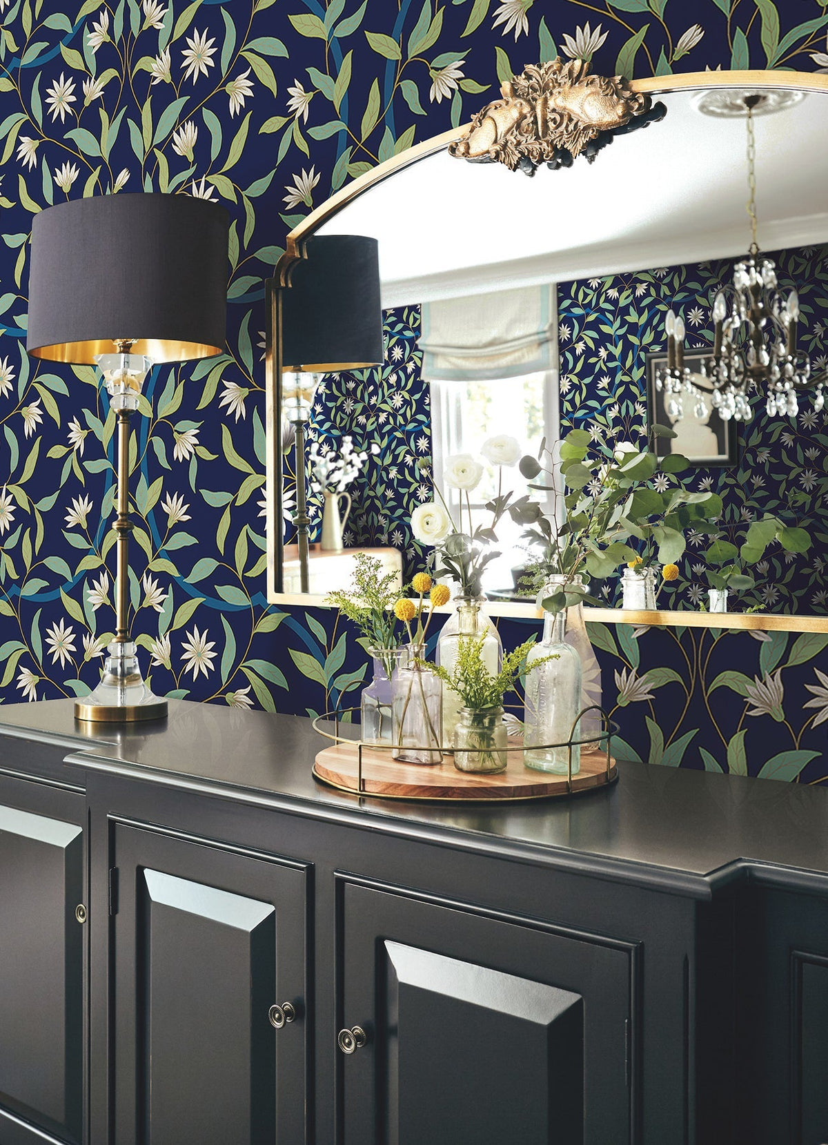 A dark cabinet with a gold tray holding vases and greenery sits below a large mirror, reflecting the Jasmine Wallpaper by York Wallcoverings and a lamp with a black shade in a stylish, elegant room.