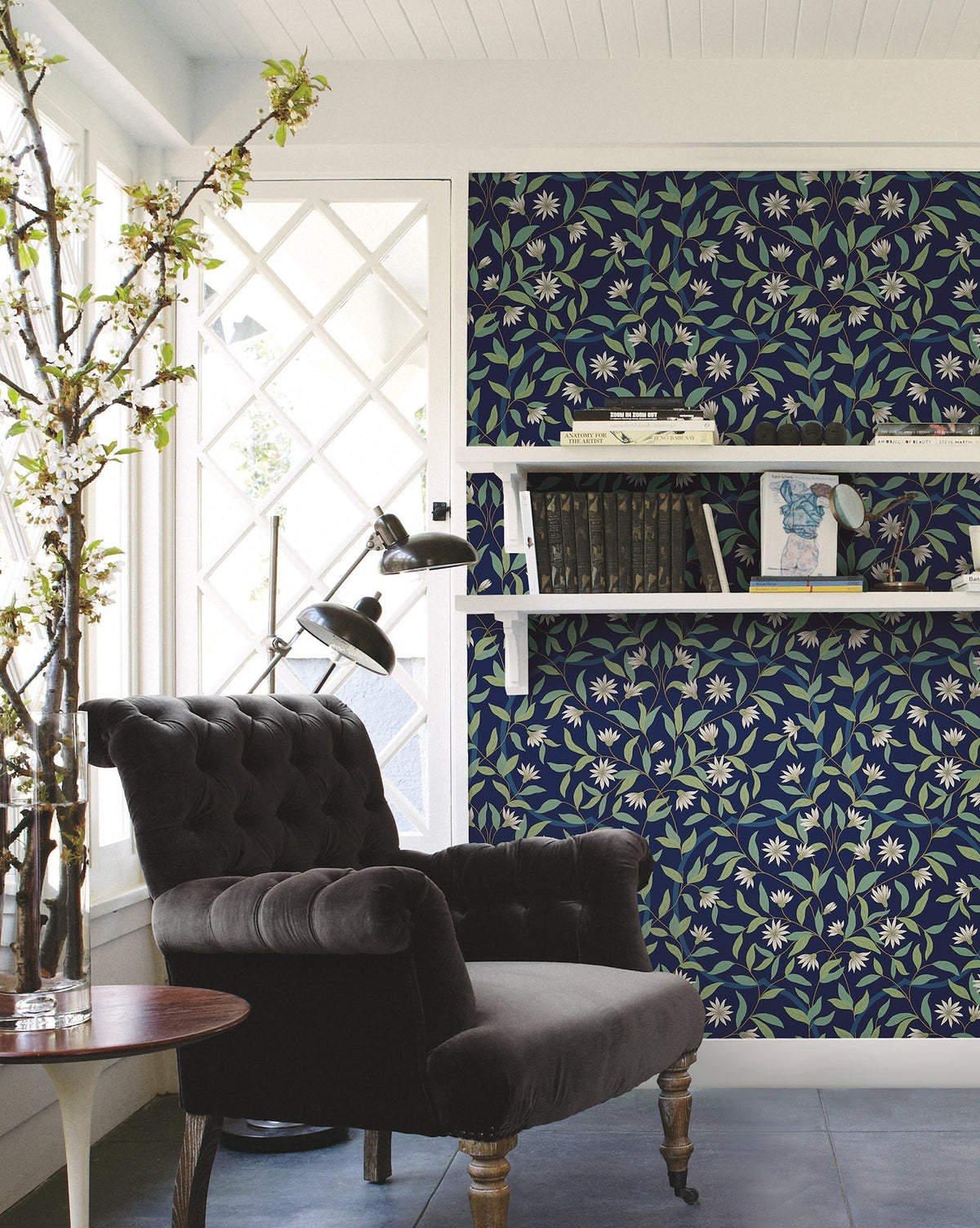 A cozy reading nook with a black tufted armchair, a round wooden side table, and a white bookshelf set against a wall covered in Jasmine Wallpaper by York Wallcoverings near large windows with white lattice trim.
