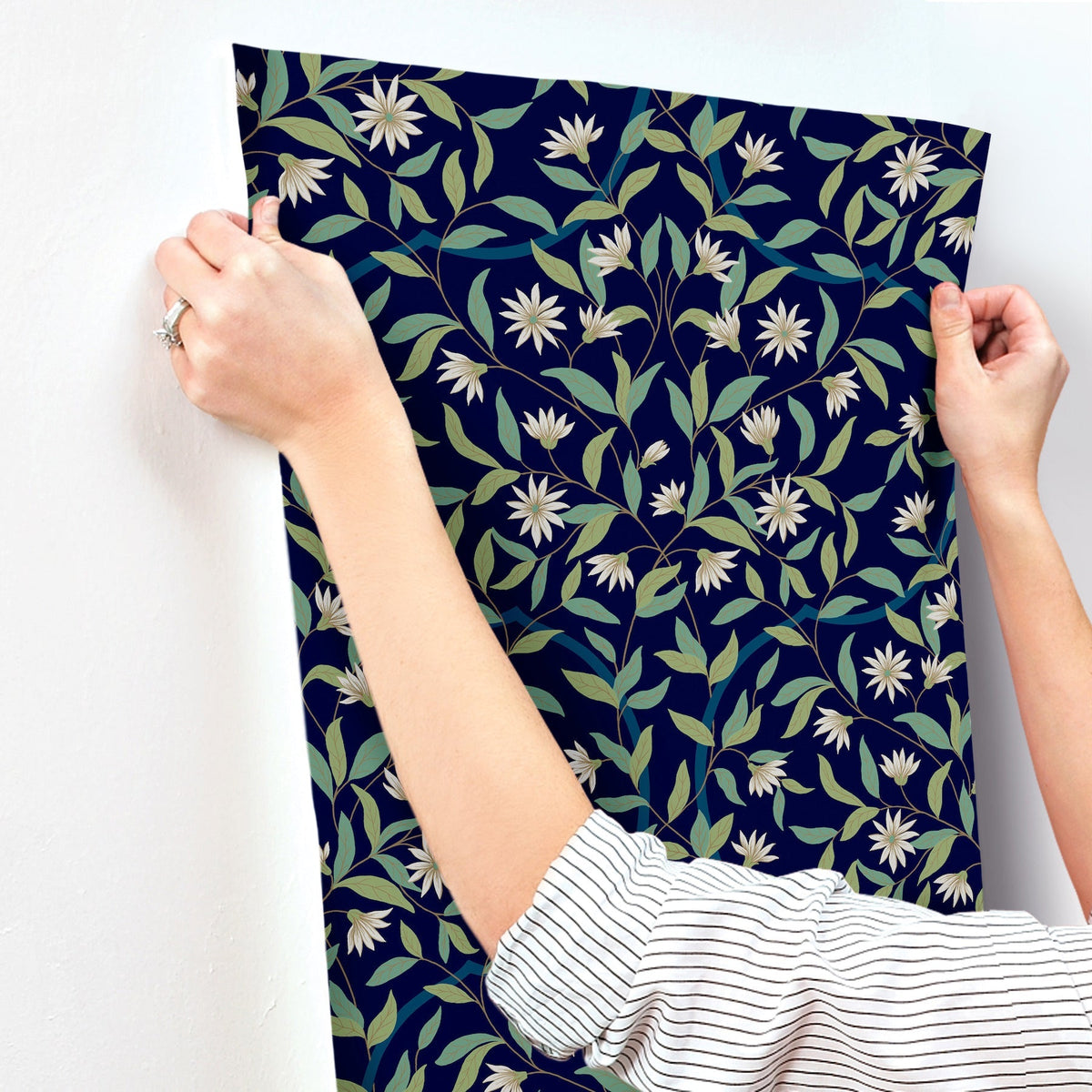A person in a striped shirt presses York Wallcoverings’ Jasmine Wallpaper onto a white wall, adjusting it with both hands.