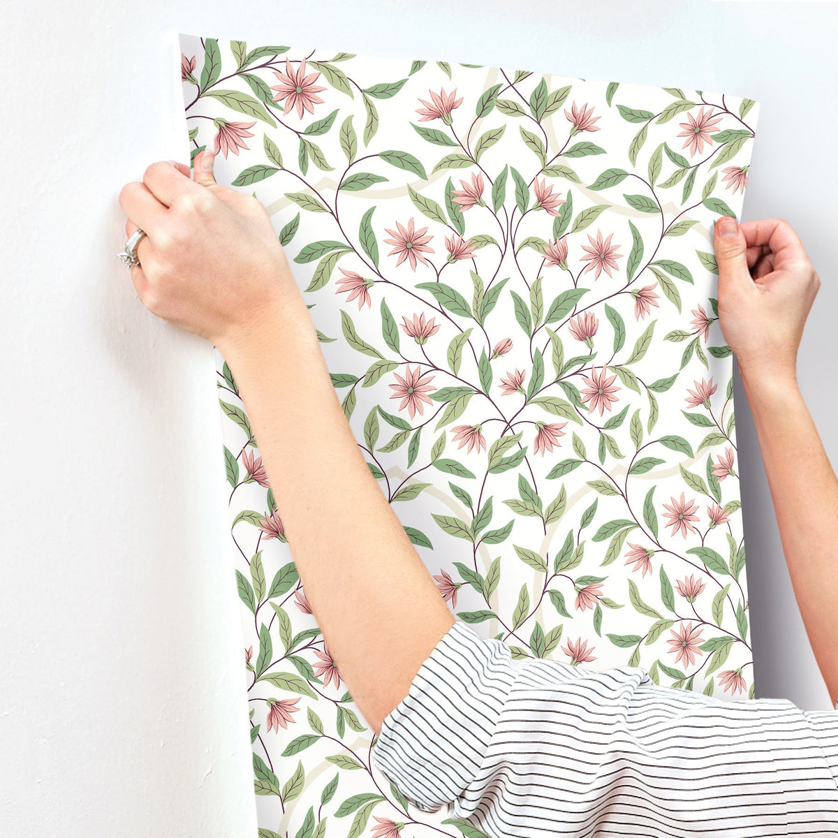 Wearing a striped sleeve, a person aligns York Wallcoverings Jasmine Wallpaper—featuring pink flowers and green leaves—against a white wall in preparation for hanging.