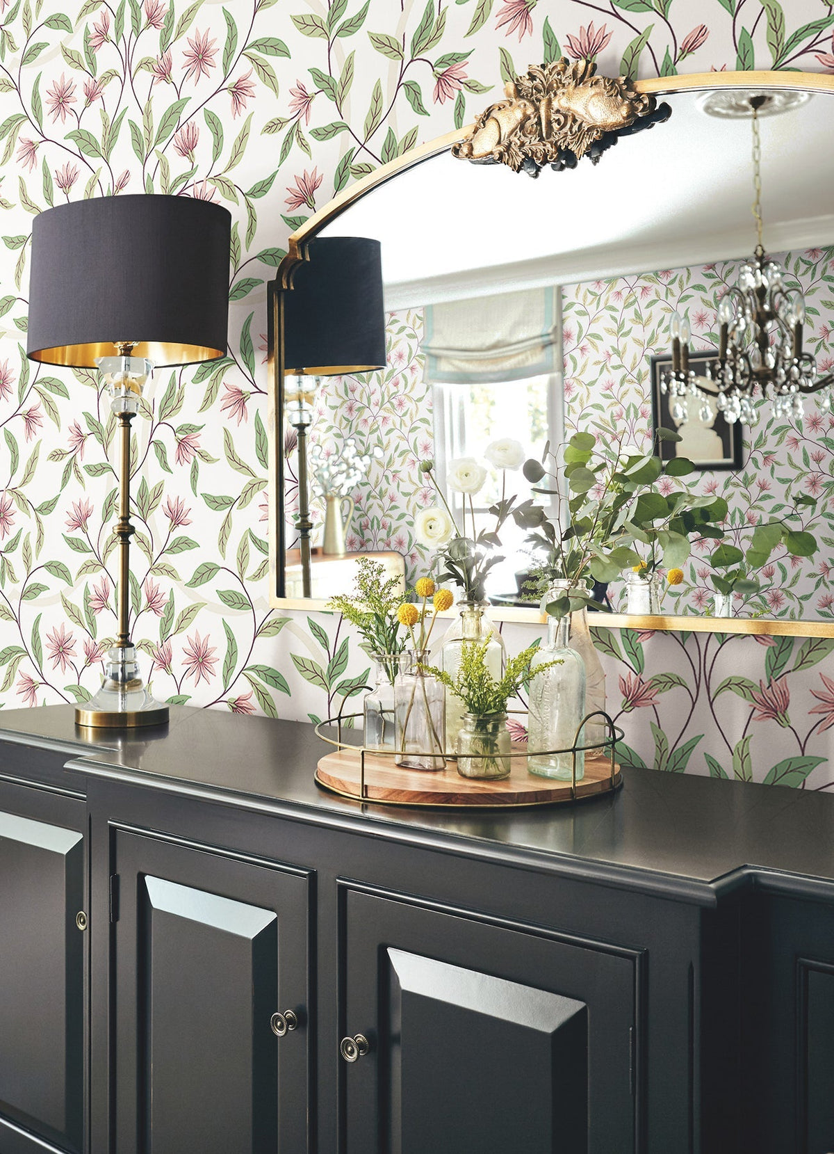 A black sideboard with glass vases, greenery, and a tray sits below a large mirror. The wall is adorned with Jasmine Wallpaper by York Wallcoverings, while a lamp with a black shade stands to the left. A chandelier is reflected in the mirror.