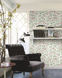 A cozy reading nook featuring a black tufted armchair, floor lamp, round side table, and shelves displayed on a Jasmine Wallpaper by York Wallcoverings, situated next to a window with white framing and a blossoming branch in a vase.