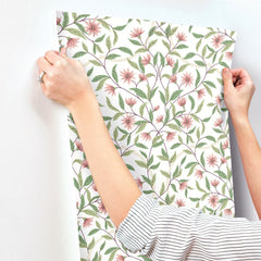 A person in a striped shirt is applying York Wallcoverings’ Jasmine Wallpaper, which features pink flowers and green leaves, onto a white wall.