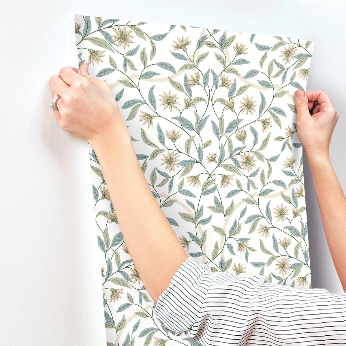 Wearing a striped shirt, a person hangs York Wallcoverings’ Jasmine Wallpaper—featuring green leaves and yellow flowers—on a white wall.