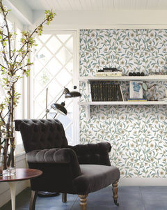 A cozy reading nook with a black armchair, side table, and floor lamp is set by a window. Jasmine Wallpaper by York Wallcoverings creates a floral backdrop, while shelves with books and a branch in a vase add charm to the space.