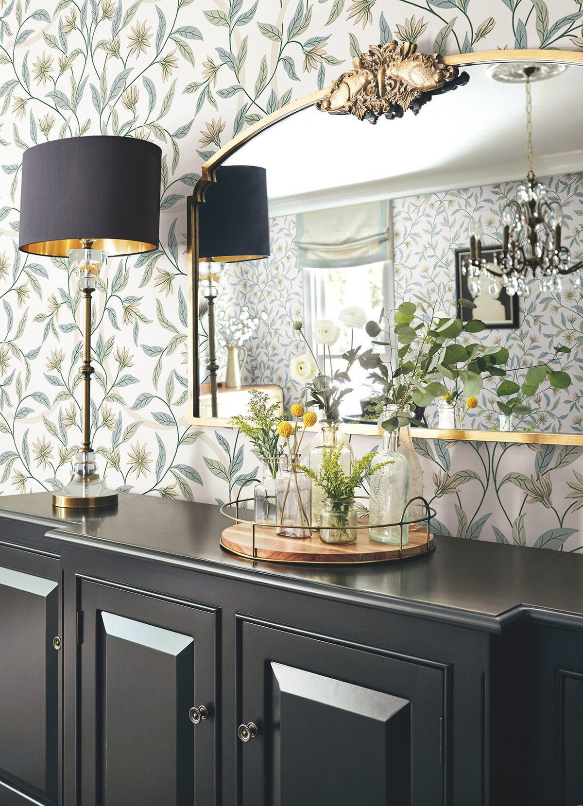 A black sideboard with a tray of glass vases, a tall black-and-gold lamp, and a large mirror sits against Jasmine Wallpaper by York Wallcoverings; a chandelier and window are reflected in the mirror.
