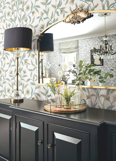 A black sideboard with a tray of glass vases, a tall black-and-gold lamp, and a large mirror sits against Jasmine Wallpaper by York Wallcoverings; a chandelier and window are reflected in the mirror.