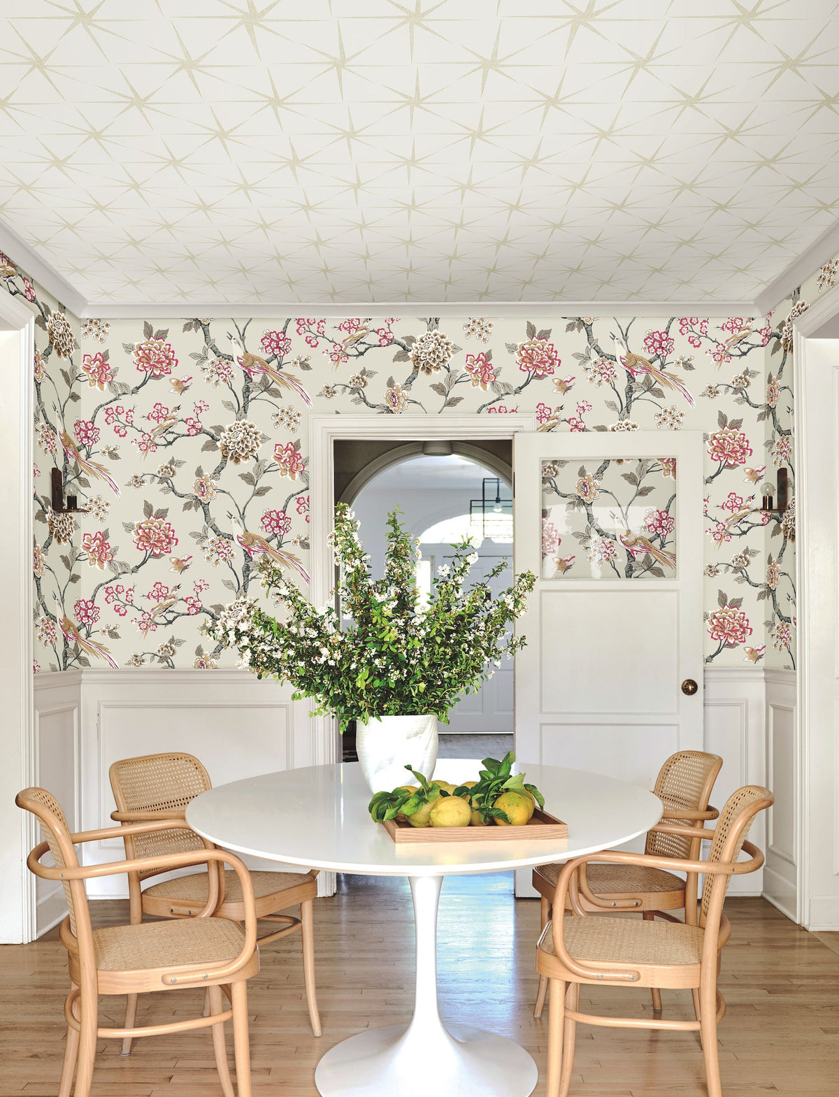 A round white table with a vase of white flowers is surrounded by four wooden chairs in a bright room featuring Fanciful Wallpaper by York Wallcoverings, light wood floors, and a bowl of lemons on the table.