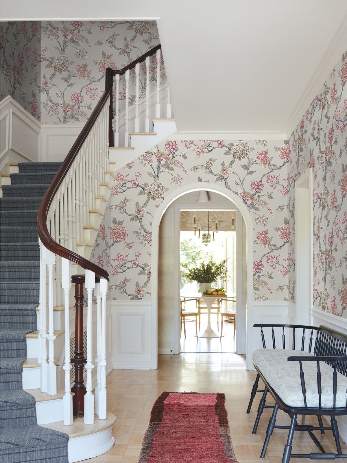 A bright hallway features Fanciful Wallpaper by York Wallcoverings, a curved staircase with a dark railing, a wooden bench, a red rug, and an arched doorway leading to a room with a dining table and chairs.