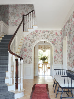 A bright hallway features Fanciful Wallpaper by York Wallcoverings, a curved staircase with a dark railing, a wooden bench, a red rug, and an arched doorway leading to a room with a dining table and chairs.