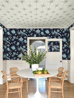 A dining room featuring a round white table, four wicker chairs, “Fanciful Wallpaper” by York Wallcoverings with a floral motif, a vase of branches, lemons on the table, and geometric patterns on the ceiling. The space also includes white doors and wood flooring.