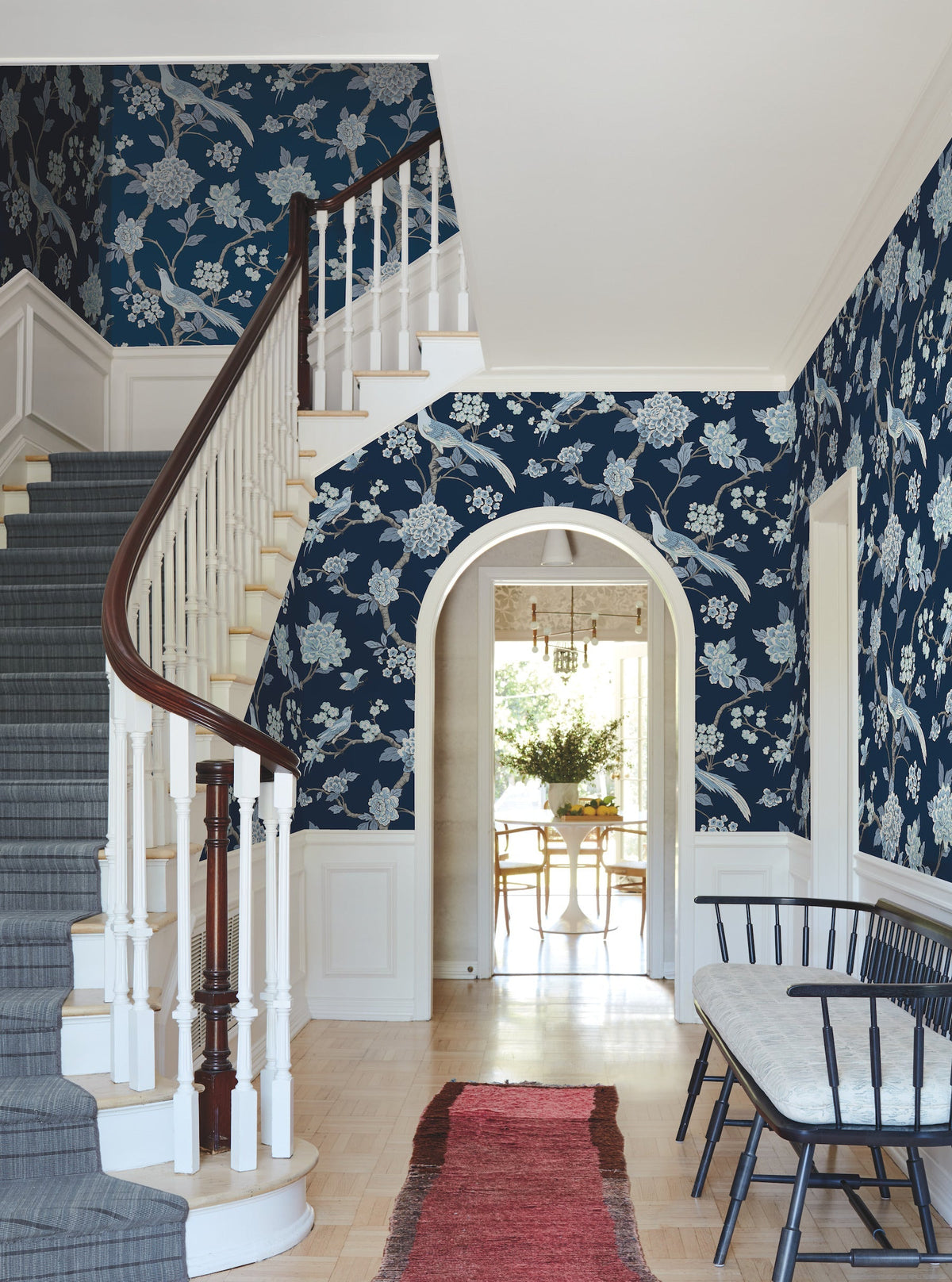 A hallway adorned with York Wallcoverings’ Fanciful Wallpaper, featuring blue florals, a curved staircase with a dark wood railing, a red rug, a black bench, and an arched doorway leading to a dining area with a table and greenery.