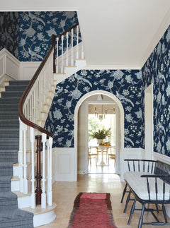 A hallway adorned with York Wallcoverings’ Fanciful Wallpaper, featuring blue florals, a curved staircase with a dark wood railing, a red rug, a black bench, and an arched doorway leading to a dining area with a table and greenery.
