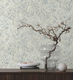 Ronald Redding Aslan Wallpaper featuring an intricate pattern of overlapping blue and grey petal-like shapes, displayed behind a shelf with a clear vase, a bowl of small stones, and a decorative orb.