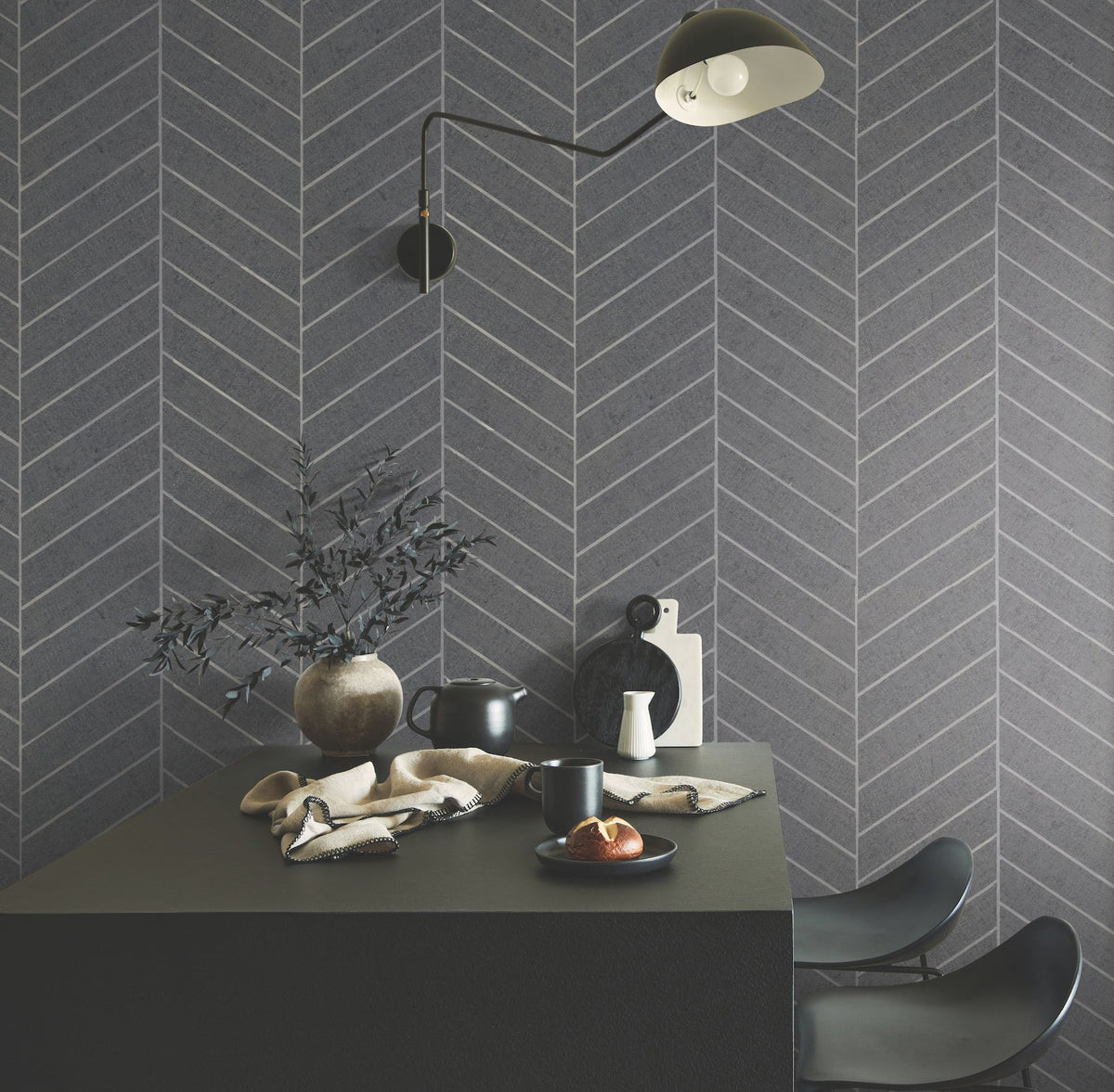 Modern dining area featuring a dark table, two black chairs, a vase with branches, cups, a croissant on a plate, and Ronald Redding Atelier Herringbone Wallpaper by York Wallcoverings in gray tones with a black wall lamp overhead.