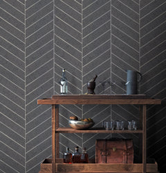 A wooden bar cart displays bottles, a bowl of fruit, glasses, a pitcher, and a leather bag, set against a dark gray wall adorned with Ronald Redding Atelier Herringbone Wallpaper by York Wallcoverings.