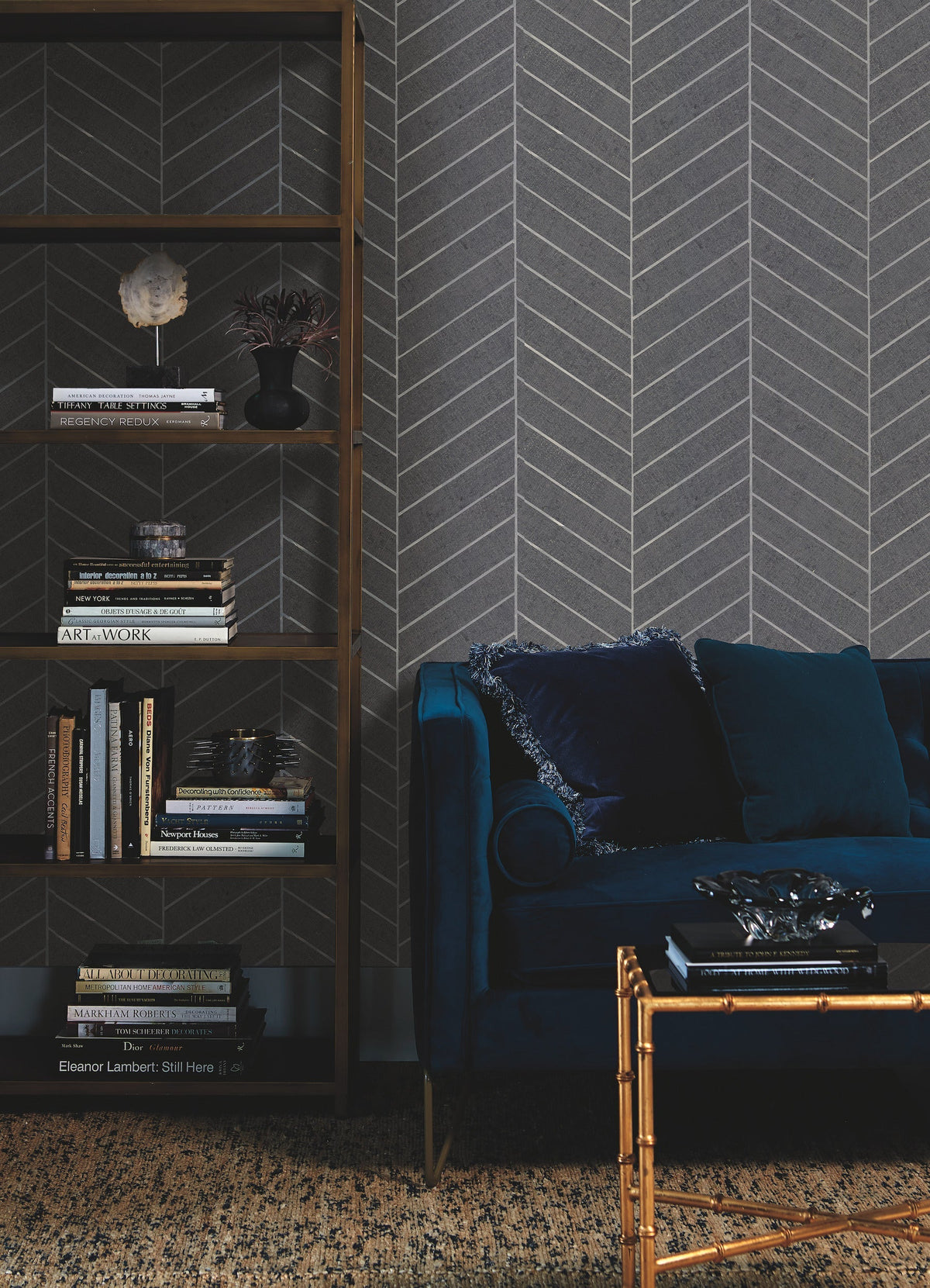 A dark velvet sofa with a blue pillow sits beside a tall bookshelf filled with books and decor, set against a gray Ronald Redding Atelier Herringbone Wallpaper by York Wallcoverings and a textured rug.