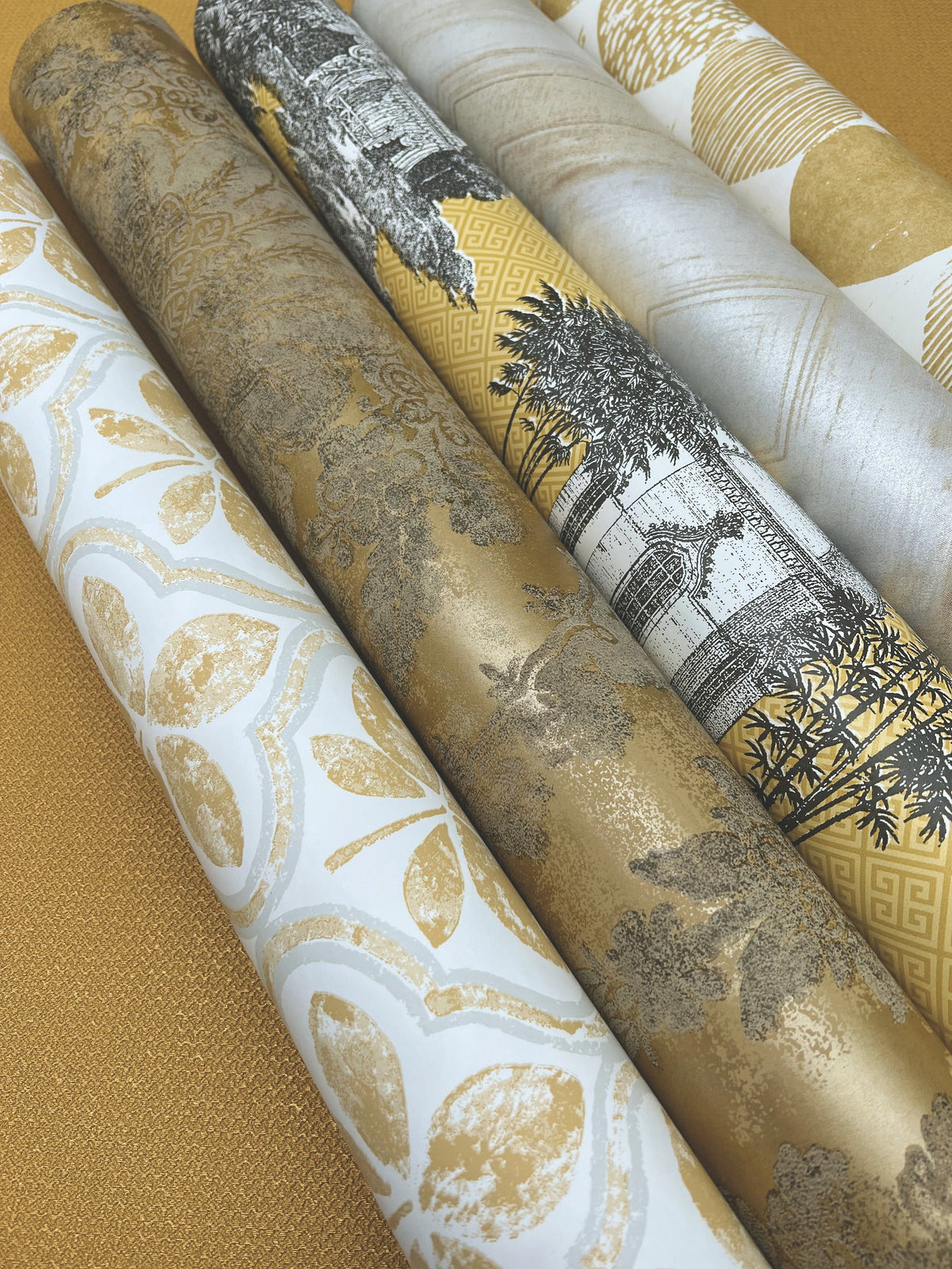 Five rolls of Ronald Redding Charleston Damask Wallpaper by York Wallcoverings featuring various botanical and architectural patterns in gold, white, and black tones.
