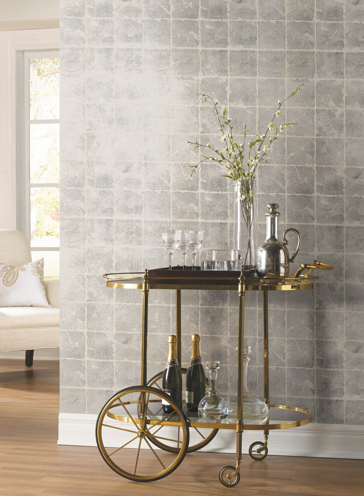 A gold bar cart adorned with glassware, decanters, and bottles sits against a wall accented with Ronald Redding Metal Leaf Squares Wallpaper by York Wallcoverings. In the background, a tall vase filled with simple branches and a window beside a white chair are visible.