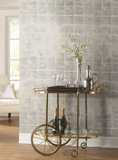 A gold bar cart adorned with glassware, decanters, and bottles sits against a wall accented with Ronald Redding Metal Leaf Squares Wallpaper by York Wallcoverings. In the background, a tall vase filled with simple branches and a window beside a white chair are visible.