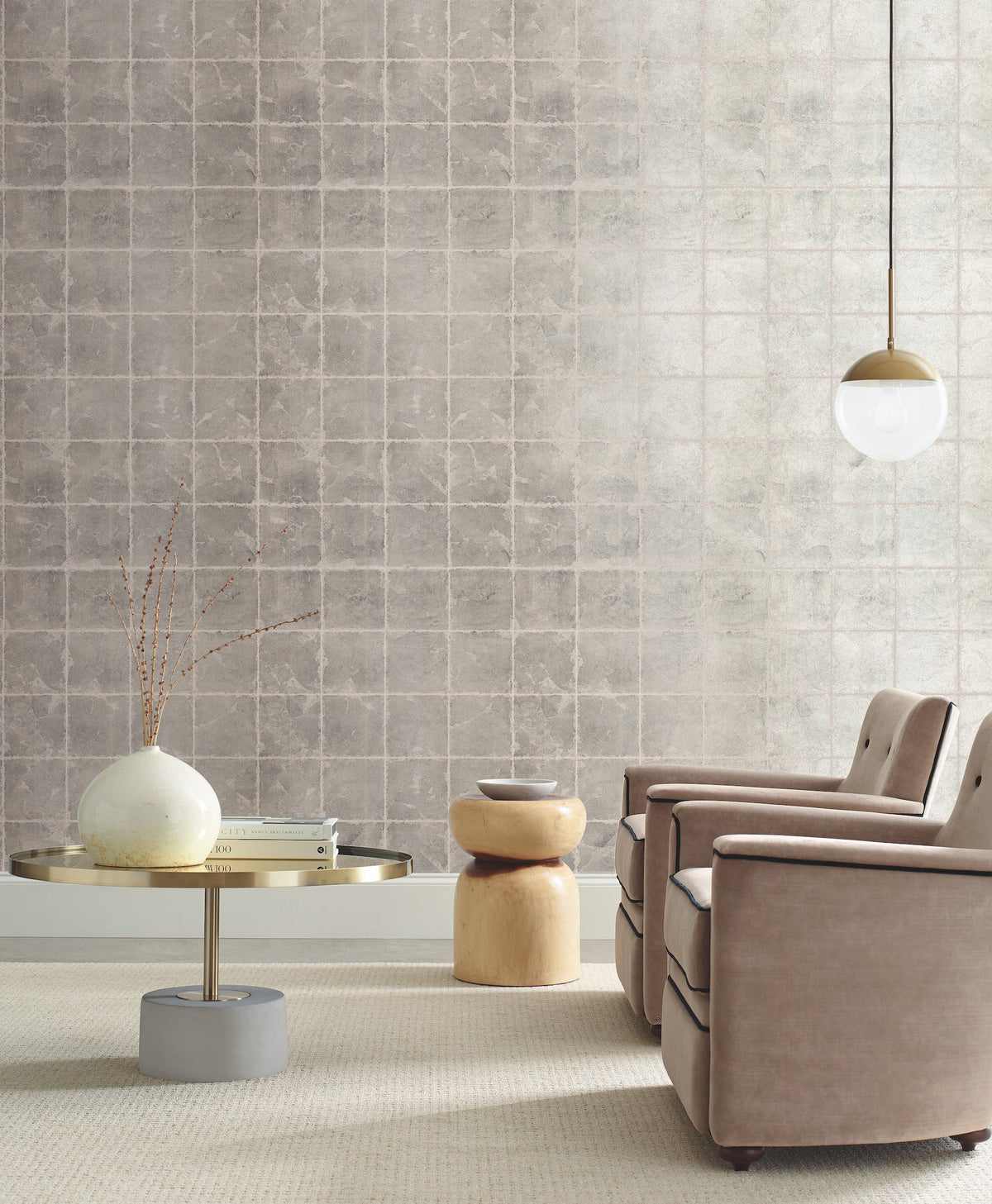 A modern living room with two beige armchairs, a round metal coffee table with a vase, a small wooden stool, and an accent wall featuring Ronald Redding Metal Leaf Squares Wallpaper by York Wallcoverings.