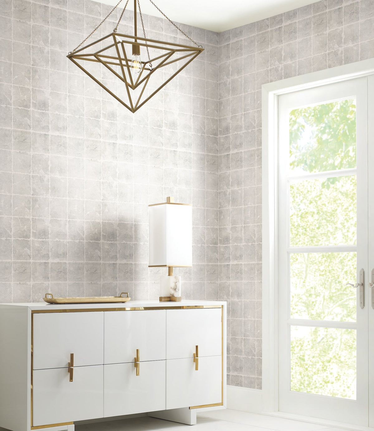 Modern room featuring a geometric brass chandelier, white sideboard with gold handles, a white lamp, and a tray. Sunlight streams through a glass door with greenery visible outside. The light gray tiled wall background is adorned with Ronald Redding Metal Leaf Squares Wallpaper by York Wallcoverings.