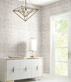 Modern room featuring a geometric brass chandelier, white sideboard with gold handles, a white lamp, and a tray. Sunlight streams through a glass door with greenery visible outside. The light gray tiled wall background is adorned with Ronald Redding Metal Leaf Squares Wallpaper by York Wallcoverings.
