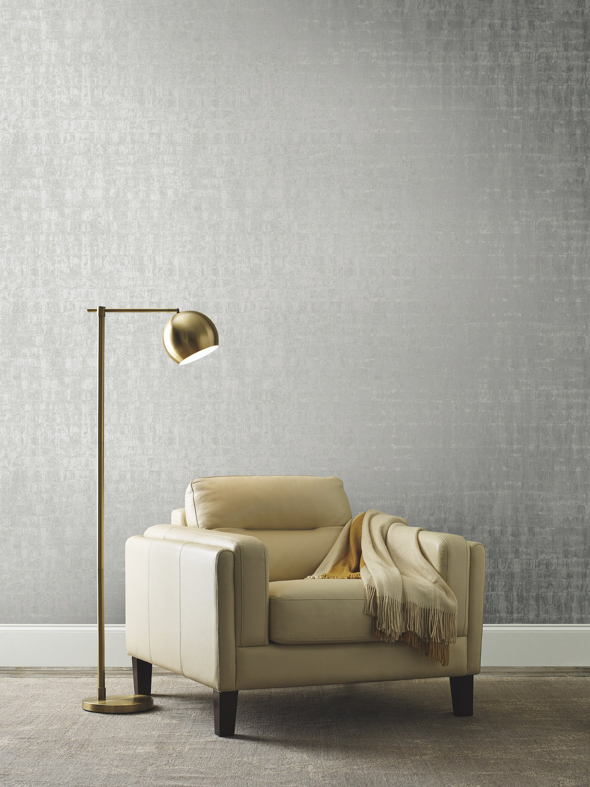 An armchair with a throw blanket and a floor lamp positioned in front of a wall featuring Ronald Redding Liquid Metal Wallpaper with a textured silver finish.
