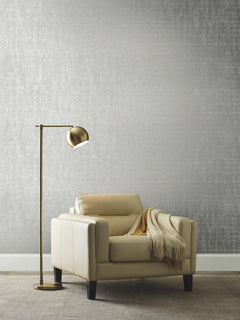 An armchair with a throw blanket and a floor lamp positioned in front of a wall featuring Ronald Redding Liquid Metal Wallpaper with a textured silver finish.