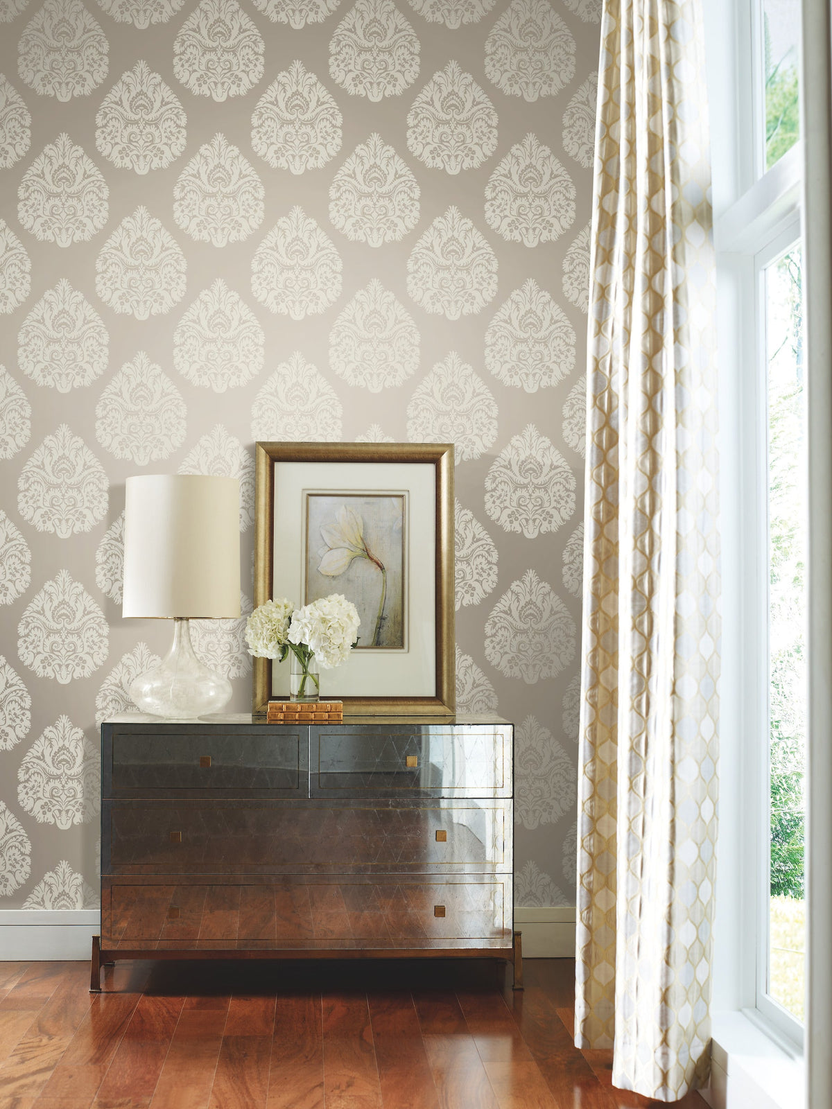 Ronald Redding Teardrop Damask Wallpaper in light gray with a repeating white teardrop damask pattern applied to a wall behind a dark wooden dresser, a lamp, a framed floral print, and a vase of white flowers.