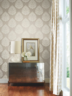 Ronald Redding Teardrop Damask Wallpaper in light gray with a repeating white teardrop damask pattern applied to a wall behind a dark wooden dresser, a lamp, a framed floral print, and a vase of white flowers.