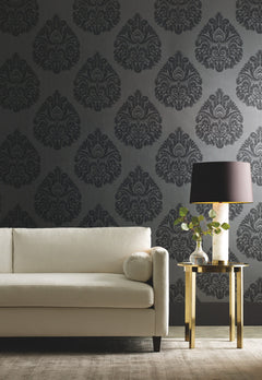 Ronald Redding Teardrop Damask Wallpaper in dark gray with a repeating teardrop pattern behind a cream-colored sofa and a gold side table with a lamp.