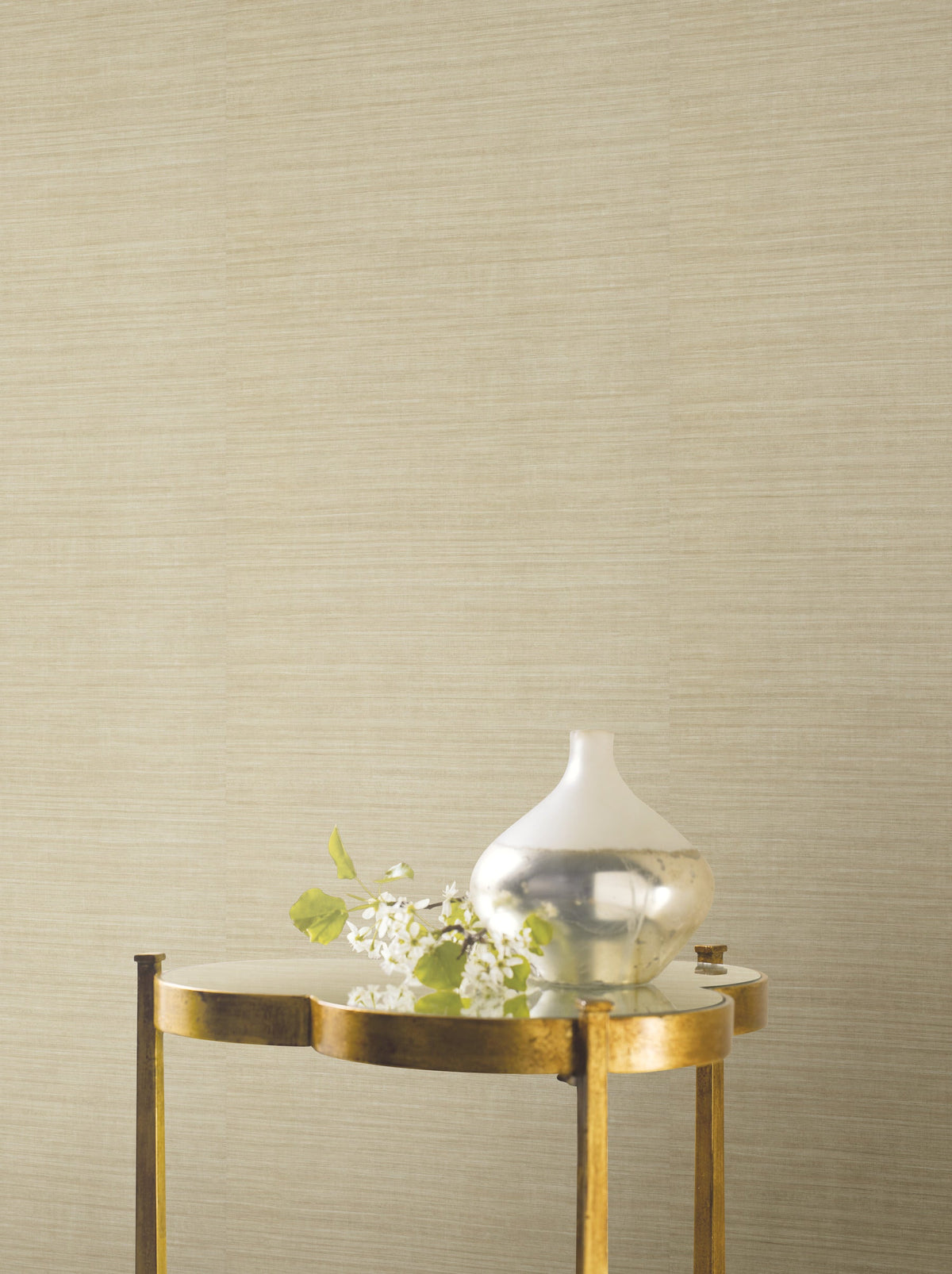 A gold side table with a mirrored top holds a small silver vase with green leaves and white flowers in front of a beige Ronald Redding Silk Elegance Wallpaper by York Wallcoverings.