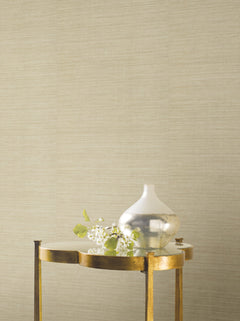 A gold side table with a mirrored top holds a small silver vase with green leaves and white flowers in front of a beige Ronald Redding Silk Elegance Wallpaper by York Wallcoverings.