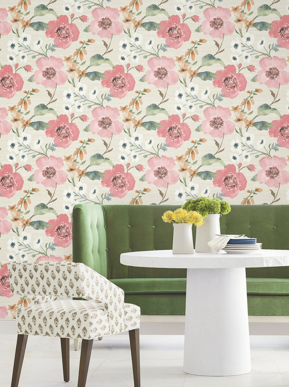 A green tufted bench and a white patterned chair surround a small white round table with yellow flowers, set against York Wallcoverings’ Evelyn Floral Wallpaper.