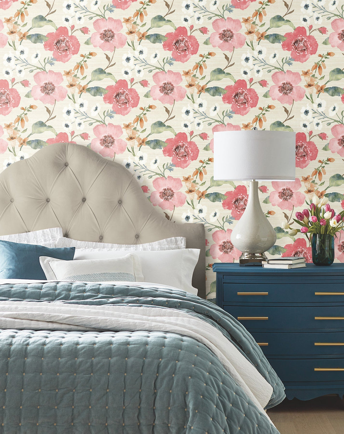 Bedroom with a tufted beige headboard, blue bedding, a navy blue dresser, lamp, and vase of tulips. The wall is adorned with Evelyn Floral Wallpaper by York Wallcoverings, featuring large pink and green flowers.