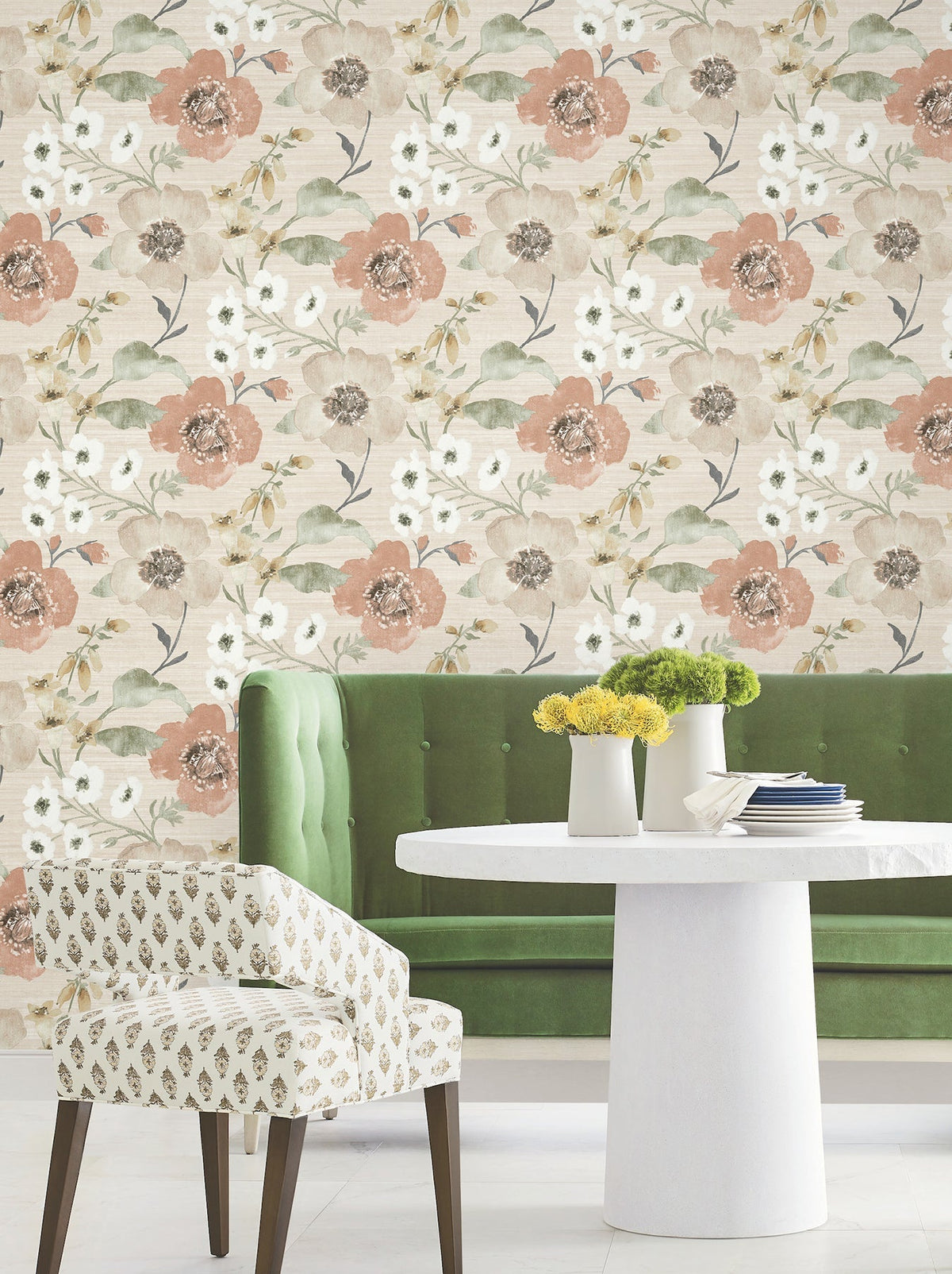 A green velvet bench and a white round table with small vases of yellow flowers sit in front of York Wallcoverings’ Evelyn Floral Wallpaper, with a patterned chair in the foreground.