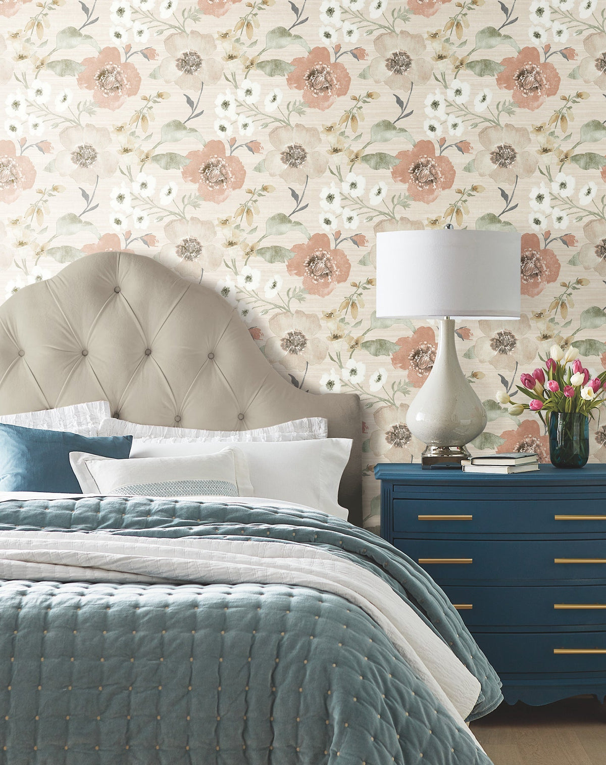 A cozy bedroom features a tufted beige headboard, blue quilted bedding, a blue dresser with gold handles, a white lamp, and a vase of pink tulips, set against Evelyn Floral Wallpaper by York Wallcoverings.