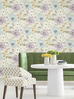 A round white table adorned with yellow flowers in vases sits beside a green tufted bench and a patterned chair, set against Evelyn Floral Wallpaper by York Wallcoverings.