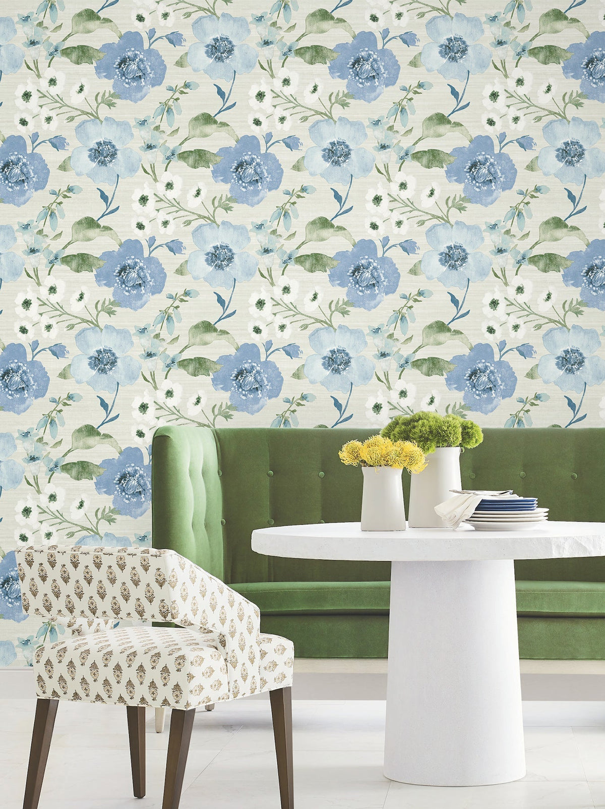A round white table adorned with yellow and green flowers, a green velvet bench, and a patterned chair are arranged in front of York Wallcoverings’ Evelyn Floral Wallpaper.