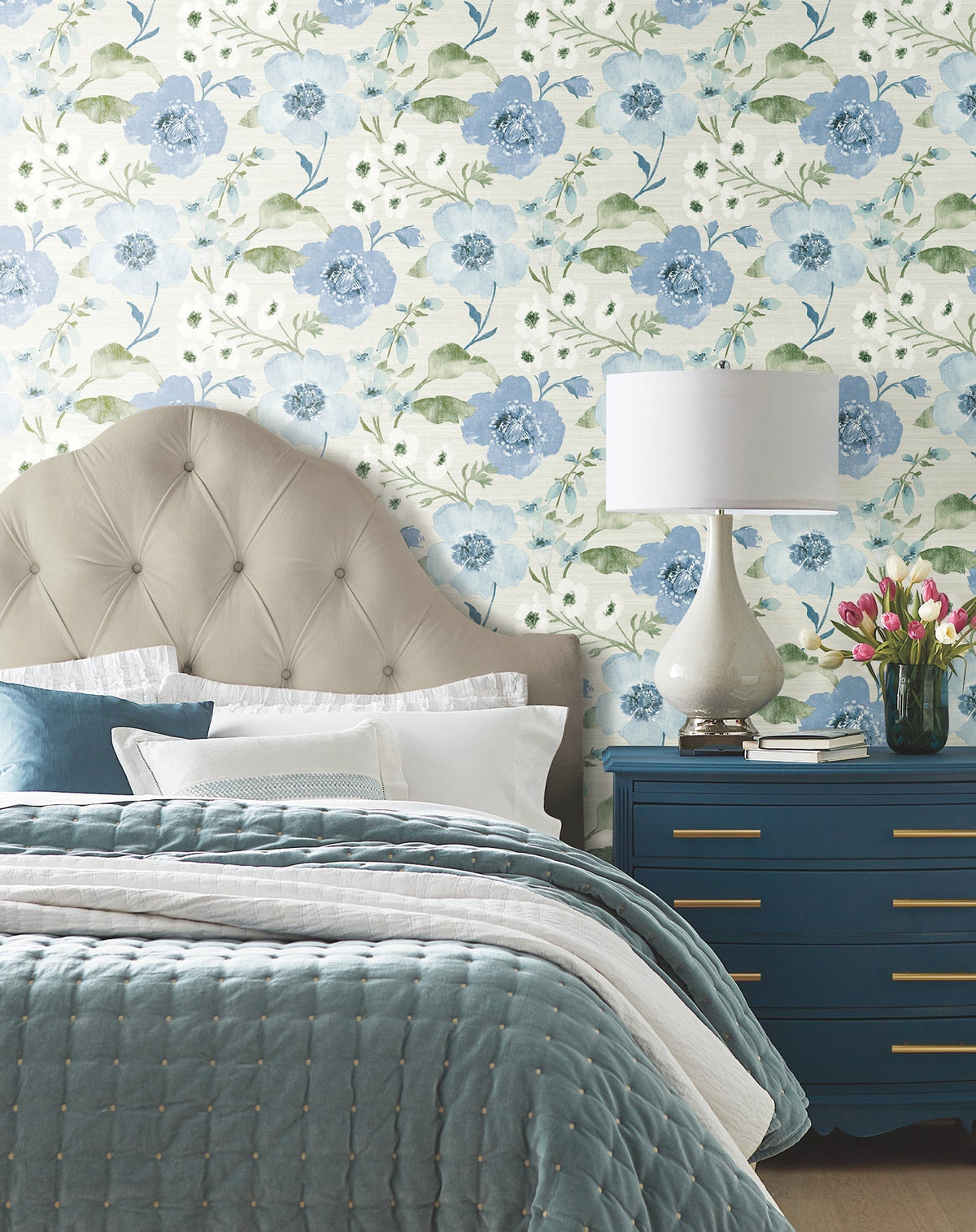 A cozy bedroom featuring a tufted beige headboard, blue and white bedding, a blue dresser, a white lamp, a vase of tulips, and Evelyn Floral Wallpaper by York Wallcoverings.
