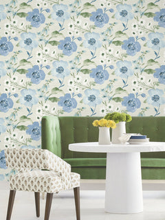 A green tufted bench, round white table, and patterned chair sit in front of a wall covered in Evelyn Floral Wallpaper by York Wallcoverings. Yellow flowers in white vases are arranged on the table.