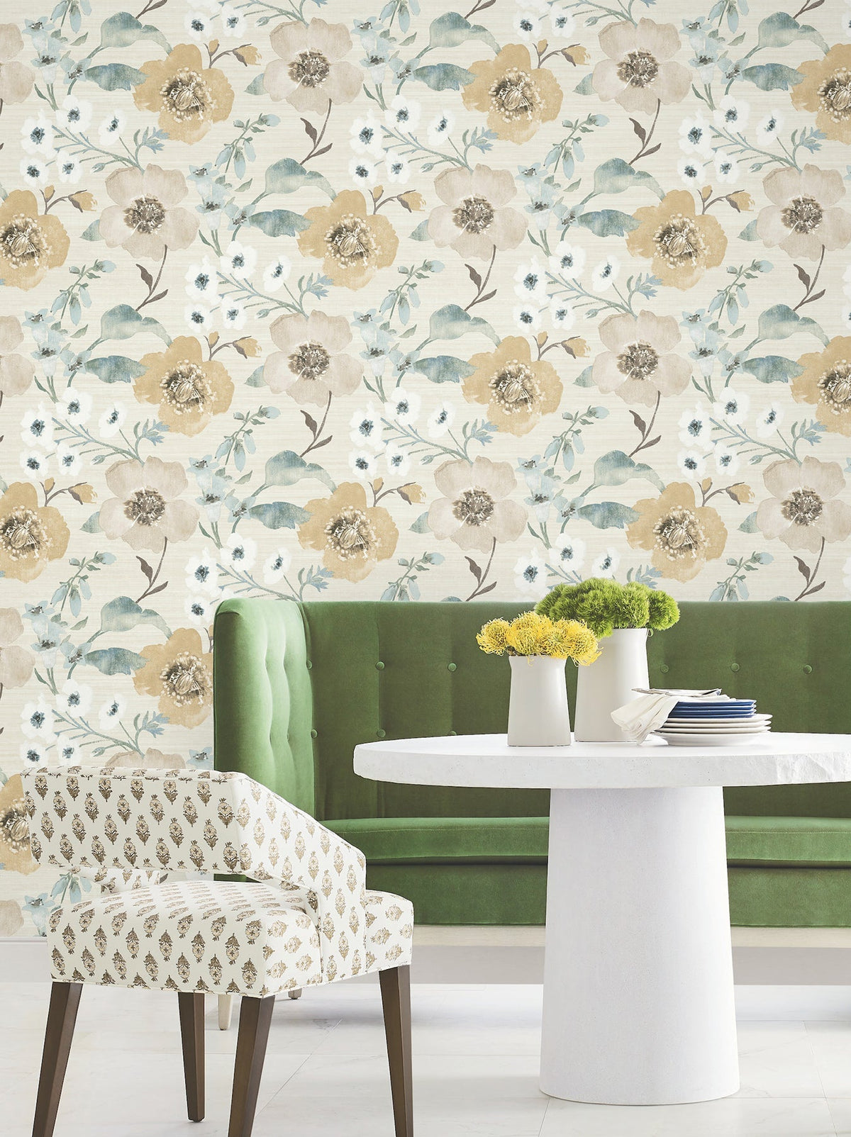 A green tufted bench, a white round table topped with vases of yellow flowers, and a patterned chair are arranged in front of the Evelyn Floral Wallpaper by York Wallcoverings.