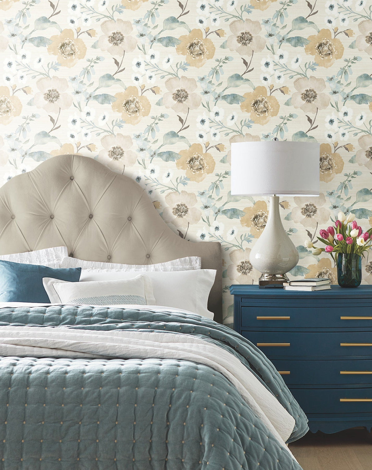 A cozy bedroom featuring a tufted beige headboard, Evelyn Floral Wallpaper by York Wallcoverings, a blue quilt, blue dresser, white lamp, and a vase of fresh tulips. White pillows and sheets brighten the bed.