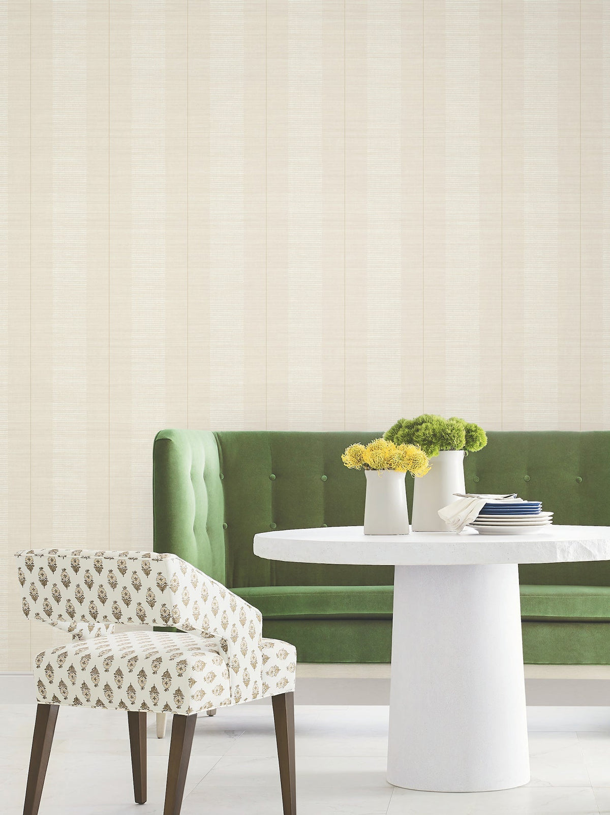 A modern dining area featuring a round white table, a green upholstered bench, a patterned chair, and vases of yellow and green flowers set against Colbert Stripe Textured Wallpaper in light beige.