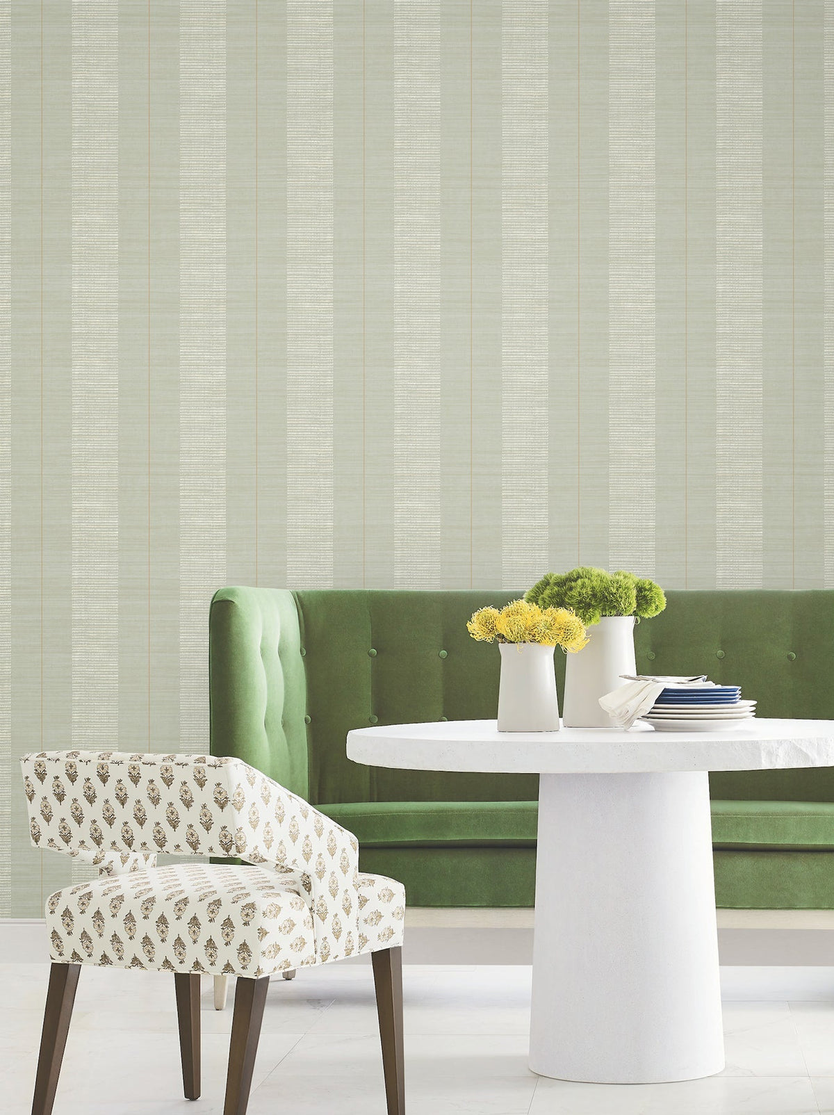 A green velvet bench, a round white table adorned with yellow and green flowers, and a white patterned chair sit against a wall covered in Colbert Stripe Textured Wallpaper.