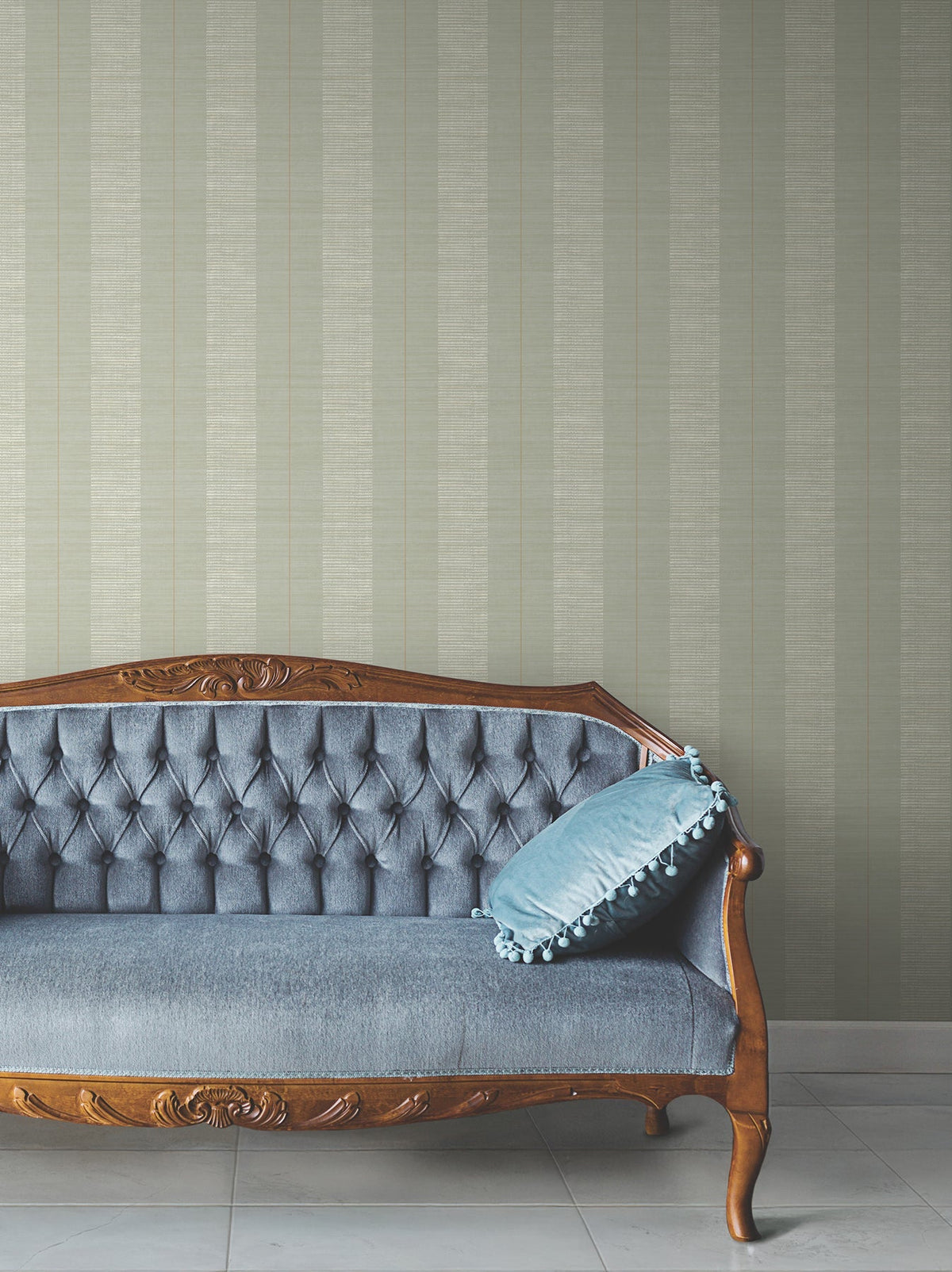 A vintage wooden sofa with blue upholstery and a decorative blue pillow sits on a tiled floor, set against a wall covered in Colbert Stripe Textured Wallpaper.