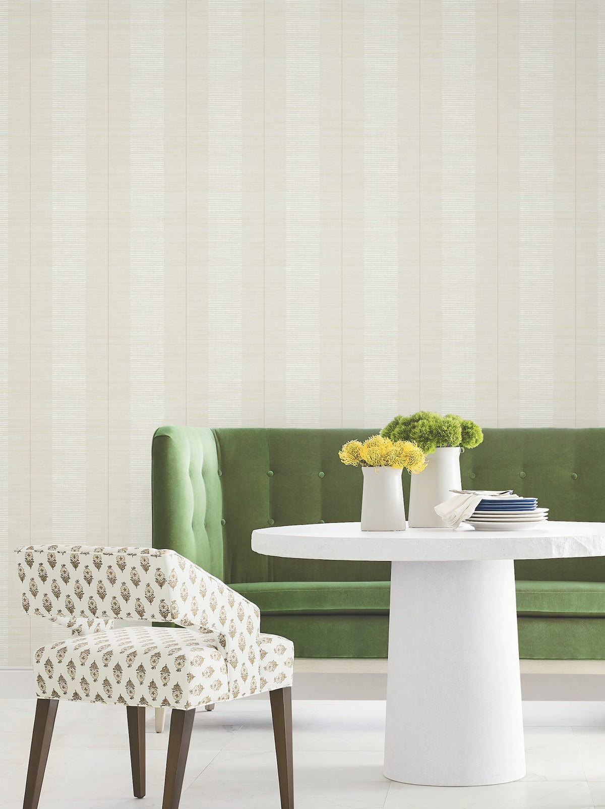 Green tufted sofa and patterned chair beside a round white table with three vases of yellow and green flowers, set against a light-colored wall adorned with Colbert Stripe Textured Wallpaper.