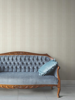 A vintage wooden sofa with blue tufted upholstery sits against a wall covered in Colbert Stripe Textured Wallpaper; a blue cushion with pom-pom trim is placed on one side of the sofa.